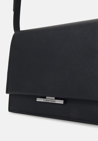 Sac bandoulière Calvin Klein en cuir texturé noir avec rabat avant et plaque logo argentée sur fond blanc.