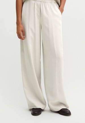 Person trägt lockere, hoch taillierte beige Hose und dunkelbraune Schuhe, mit einer Hand in der Tasche vor einfachem Hintergrund.