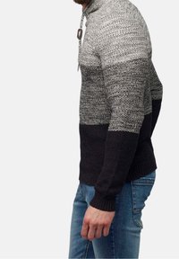 Man som bär en grå och svart stickad färgblockströja med en dragsko i kragen, tillsammans med blå jeans, sedd från sidan.