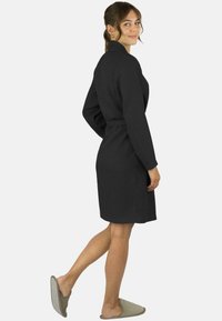 Schwarzer Bademantel mit Waffelstruktur, langen Ärmeln und Gürtel in der Taille, knielang, mit hohem Kragen und passenden hellgrauen Hausschuhen.