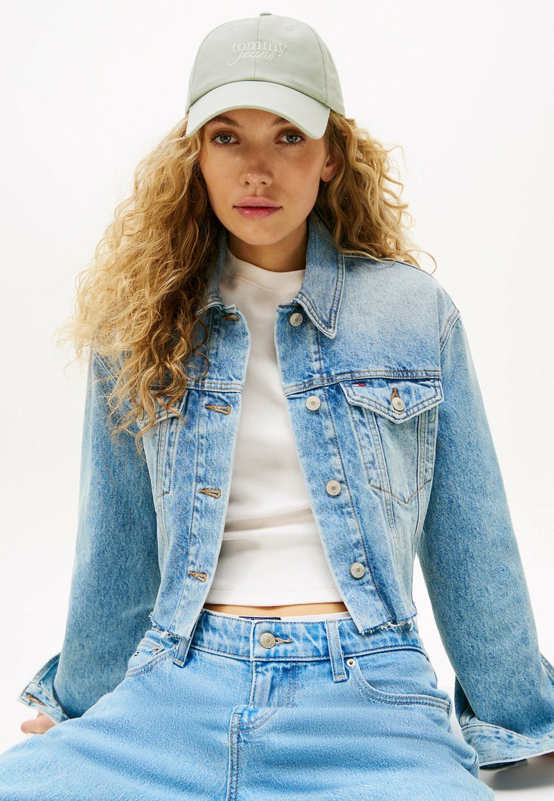 Chaqueta de mezclilla azul clara con un diseño ajustado, que presenta cierres de botón y bolsillos en el pecho, combinada con una camiseta blanca ajustada y jeans azul claros.