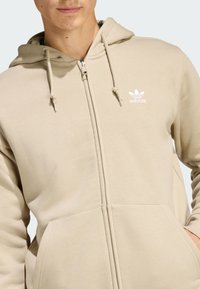 Béžová mikina na zips s kapucňou, s vreckom klokankou, nastaviteľnými šnúrkami a malým bielym logom Adidas na ľavej strane hrudníka. Jemná texturovaná látka.