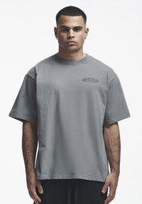 Homme adulte aux cheveux courts portant un t-shirt gris oversize avec le texte « 2Y STUDIOS » sur la poitrine, debout devant un fond blanc uni.