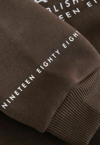 Brunt sweatshirtstof med en glat tekstur. Indeholder hvidt trykt tekst "NINETEEN EIGHTY" langs manchet og krop i en moderne skrifttype.