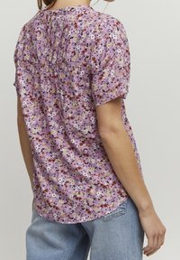 Blomstrende kortærmet top i lavendel, med en samlet ryg, flerfarvet blomstermønster og en rund kant. Stoffet synes let.