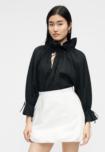 Γυναίκα με μαύρα μακριά μαλλιά, που φοράει μια μαύρη μπλούζα με βολάν και λεπτομέρειες κορδονιών και μια λευκή ψηλόμεση φούστα, σε ουδέτερο φόντο.