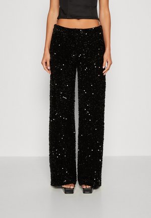 Pantalones negros de pierna ancha y talle alto cubiertos de lentejuelas negras, combinados con un top negro corto y sandalias negras de plataforma.