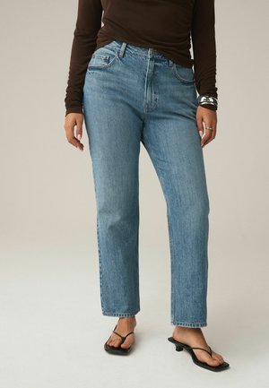 Frau trägt hellblaue Jeans mit geradem Bein, schwarze Riemchen-Sandaletten mit Absatz, ein dunkelbraunes Langarmoberteil, ein silbernes Armband und einen Ring.