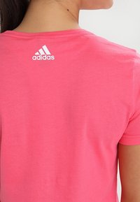 Růžové tričko s krátkým rukávem vyrobené z měkké tkaniny, s bílým logem Adidas na horní části zad. Jednoduchý design s kulatým výstřihem.