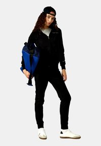 Schwarze Zip-Jacke über einem grauen Shirt, schwarze Jogginghosen, weiße Sneakers und eine blaue Sporttasche mit schwarzen Trägern, vor einem grauen Hintergrund.