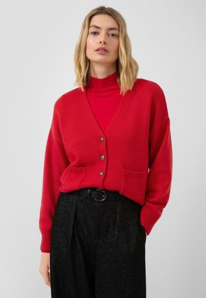 Rode gebreide cardigan met een V-hals, knoopsluiting en twee voorkruiszakken. Gecombineerd met een rode coltrui en zwarte, gestructureerde broeken.