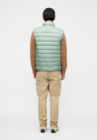 Αμάνικο μπουφάν σε ανοιχτό πράσινο μέντας, με κάθετο κεντητό μοτίβο. Φορεμένο πάνω από καφέ πλεκτό πουλόβερ, συνδυασμένο με μπεζ παντελόνι cargo.