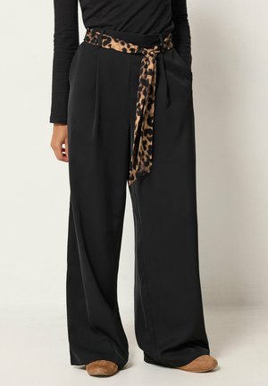 Modèle portant un pantalon noir à jambes larges, une ceinture-écharpe à imprimé léopard et des chaussures couleur fauve.