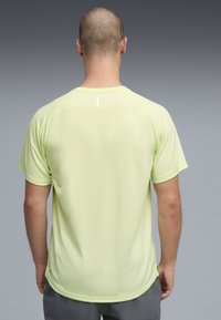 Άνδρας με ξυρισμένο κεφάλι, φορώντας αθλητική κοντομάνικη μπλούζα ανοιχτού κίτρινου χρώματος και γκρι σορτς, στέκεται με την πλάτη προς την κάμερα μπροστά σε γκρι φόντο.