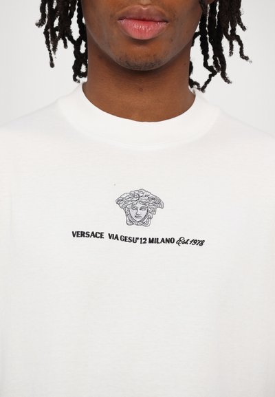 Balts kokvilnas T-krekls ar melnu izšūtu grafiku, kurā attēlota Medūzas galva un uzraksts: "VERSACE VIA JESU'12 MILANO Est. 1978."
