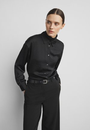 Vrouw in zwart overhemd met knopen en zwarte broek, handen in de zakken, kijkt iets opzij tegen een effen grijze achtergrond.