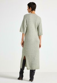 Vestito lungo verde morbido a maglia con maniche corte, spacchi laterali e vestibilità ampia. Abbinato a stivali alti di pelle nera.