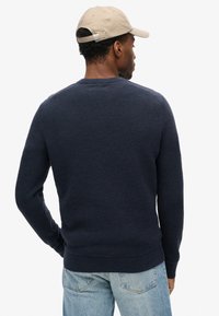 Marinefarbener Strickpullover mit strukturiertem Muster, Rundhalsausschnitt und gerippten Bündchen. Trägt ihn mit hellblauen Jeans und einer beigen Mütze.