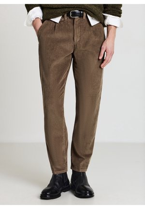 Uomo che indossa pantaloni di velluto a coste marroni, cintura nera, stivali in pelle neri, camicia bianca e maglione verde scuro con maniche risvoltate.