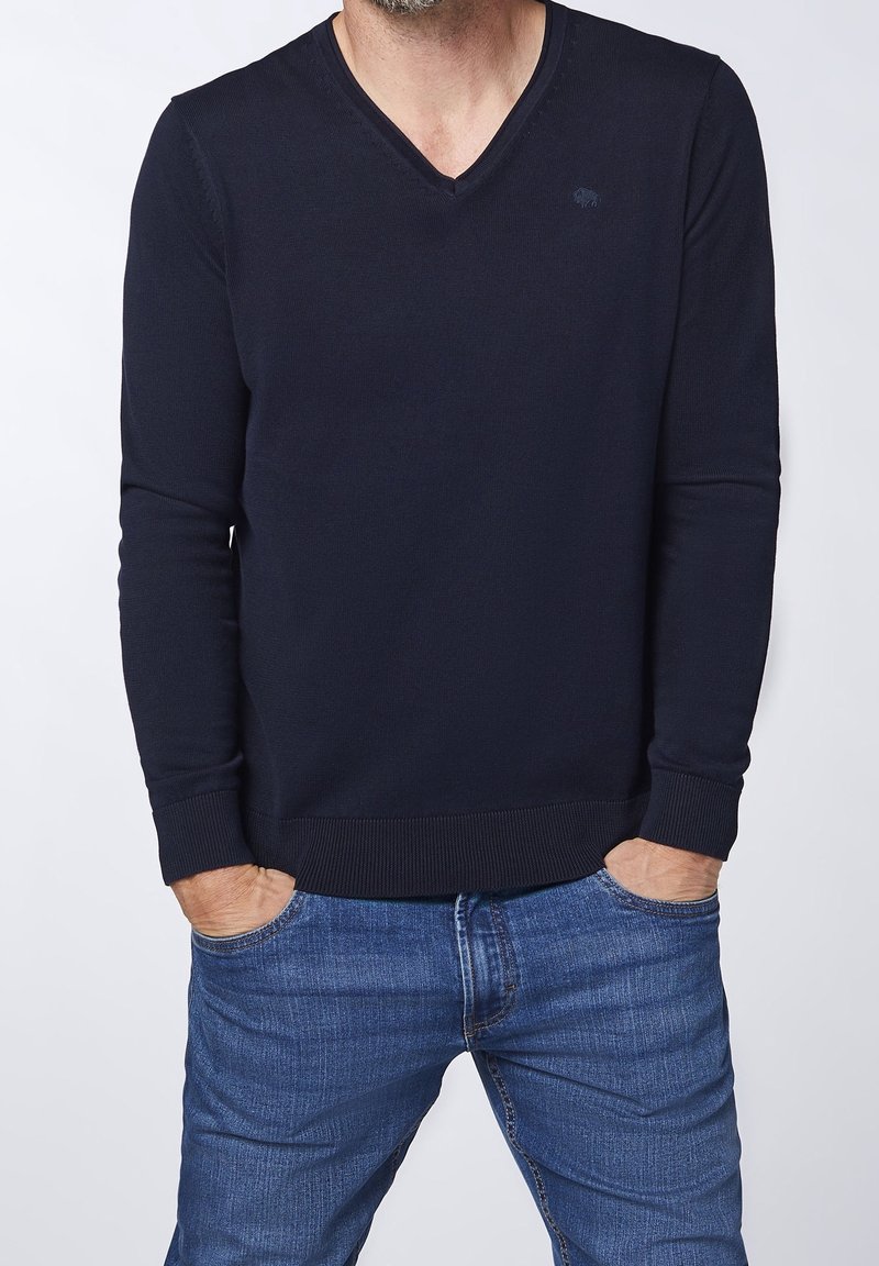 Marinblå v-ringad tröja med långa ärmar, ribbade ärmslut och nederkant, tillverkad av ett texturerat material. Liten logotyp på övre vänstra sidan.