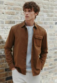 Hnedá košeľa na gombíky s vreckom na hrudi, preložená cez svetlý štruktúrovaný henley. Nastavené na pozadí s murovanou stenou, ktorá zdôrazňuje prírodné textúry.