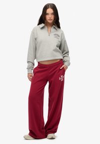 Sudadera gris recortada con cuello de cremallera y estampado de "Superdry Athletics", combinada con pantalones de chándal anchos en color burdeos con detalles del logo en blanco.