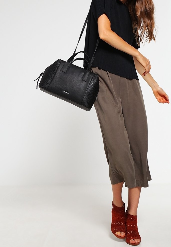 Sac à main noir structuré avec des accents texturés, deux poignées supérieures et une fermeture zippée. Associé à un pantalon olive ample et des sandales perforées rouges.