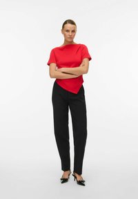 Top rojo de manga corta con un dobladillo asimétrico, combinado con pantalones negros ajustados y zapatos negros de tacón con un detalle de lazo. Diseño sencillo.