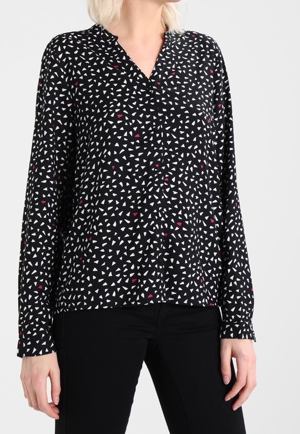 Femme portant un chemisier noir à manches longues avec un petit motif de triangles blancs et rouges, associé à un pantalon noir.