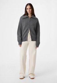 Grauer gerippter Cardigan mit durchgehendem Reißverschluss und weitem Kragen, kombiniert mit cremefarbenen, weit geschnittenen Hosen und beigen Schuhen. Einfaches Design und strukturierter Stoff.