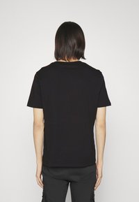 Persona con cabello oscuro hasta los hombros, vistiendo una camiseta de manga corta negra y pantalones negros, de espaldas frente a una pared blanca.
