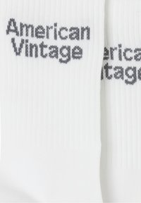 Calcetines de algodón blanco con textura acanalada, que presentan el logo "American Vintage" tejido en gris en un lado. Diseño y forma simples.