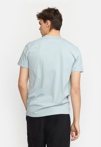 Lichtblauwe katoenen t-shirt met korte mouwen en een ronde halslijn, gezien vanuit de achterkant. Heeft een eenvoudig ontwerp en een relaxte pasvorm. Zwarte broek zichtbaar.