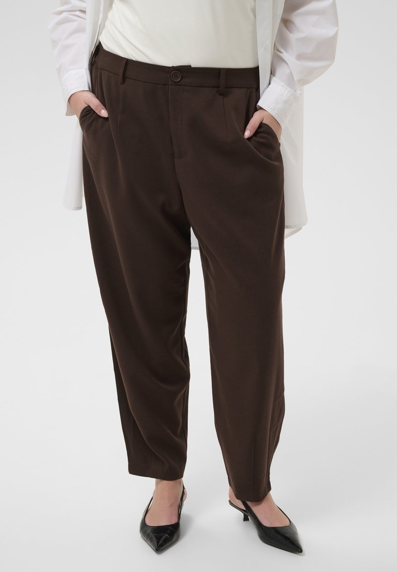 Femme portant un pantalon marron ample, un top blanc et une chemise blanche, les mains dans les poches, chaussures noires à talons bas pointus visibles.