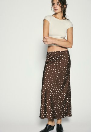 Frau trägt ein cremefarbenes kurzärmeliges Croptop und einen braunen Midirock mit Polka-Dots sowie schwarze Slipper, steht vor einer einfarbigen hellen Wand.