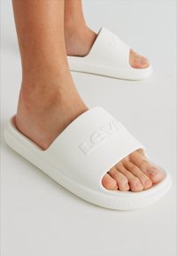 White rubber slide sandals with a textured strap, featuring an embossed logo. The sole has a ribbed pattern for grip and comfort.