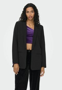 Blazer noir au design ajusté, à manches longues, fermeture à un bouton et deux poches avant. Porté par-dessus un top court violet.