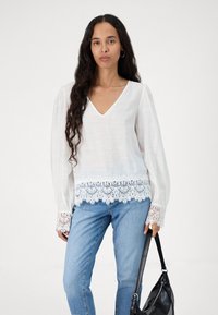 Witte blouse met lange pofmouwen, V-hals en kantdetails aan de zoom en manchetten. Gemaakt van een lichtgewicht, gestructureerde stof. Gecombineerd met blauwe jeans.