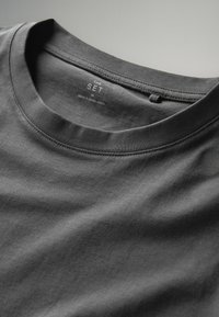 Tumšs pelēks T-krekls ar apaļu kaklu un gludu kokvilnas faktūru. Uz burtiem iekšējā apakšējā daļā rakstīts "THE SET M MADE IN BANGLADESH."