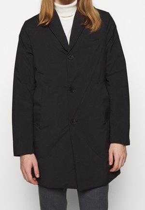 Homme portant un manteau noir boutonné sur un col roulé blanc et un pantalon gris, debout avec les mains détendues sur les côtés.