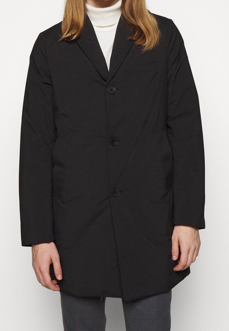 Mężczyzna w czarnym, zapinanym na guziki płaszczu, będący ubrany w biały golf i szare spodnie, stojący z rękami swobodnie opuszczonymi wzdłuż ciała.