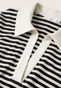 Polo a rayas en blanco y negro con textura acanalada, que presenta un cuello blanco en contraste y una abertura en forma de V. Etiqueta visible en el interior.