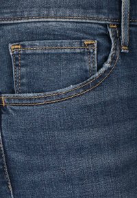 Denimjeans i en mörk indigofärg, med en bakficka som har gul söm och en något sliten textur.