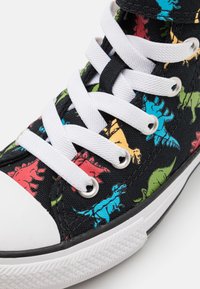 Svart canvas sneaker med färgglada dinosaurmönster, vita snören och en klassisk gummitå med strukturerad sula.