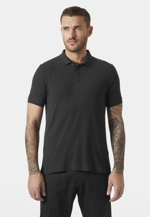 Mężczyzna z tatuażami na ramionach, ubrany w czarną koszulkę polo z krótkim rękawem i czarne spodnie, patrzący lekko na bok na białym tle.