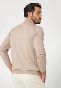 Maglione a collo alto beige con un design aderente, realizzato in un tessuto a maglia liscio. Polsini e orlo a costine, indossato con pantaloni di colore chiaro.