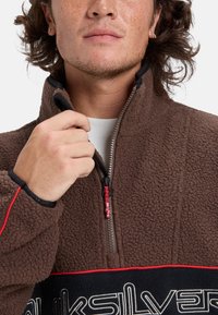 Sudadera de forro polar marrón con acentos en negro y rojo, diseño de media cremallera, textura elevada y detalle del logo en el pecho.