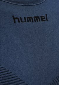 Mörkblå texturerad tyg med svart "hummel"-logotyp tryckt nära halsringningen och en subtil diagonalt ribbad struktur nedanför.
