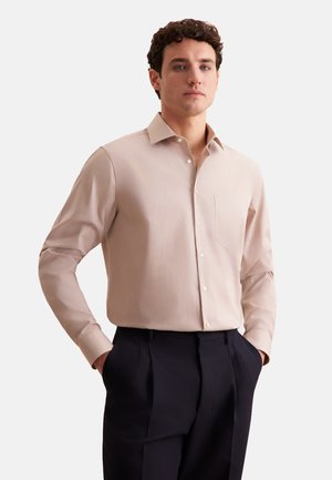 Lichtbeige langslijfshirt met een gestructureerde kraag, knoopsluiting aan de voorkant en een borstzak, gedragen bij donkere geplooide broeken. Glad textuur van de stof.