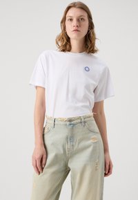 Vit bomulls-T-shirt med en blå smiley-logotyp på vänster bröst, i kombination med ljusblå denimjeans med fransar och slitna detaljer.